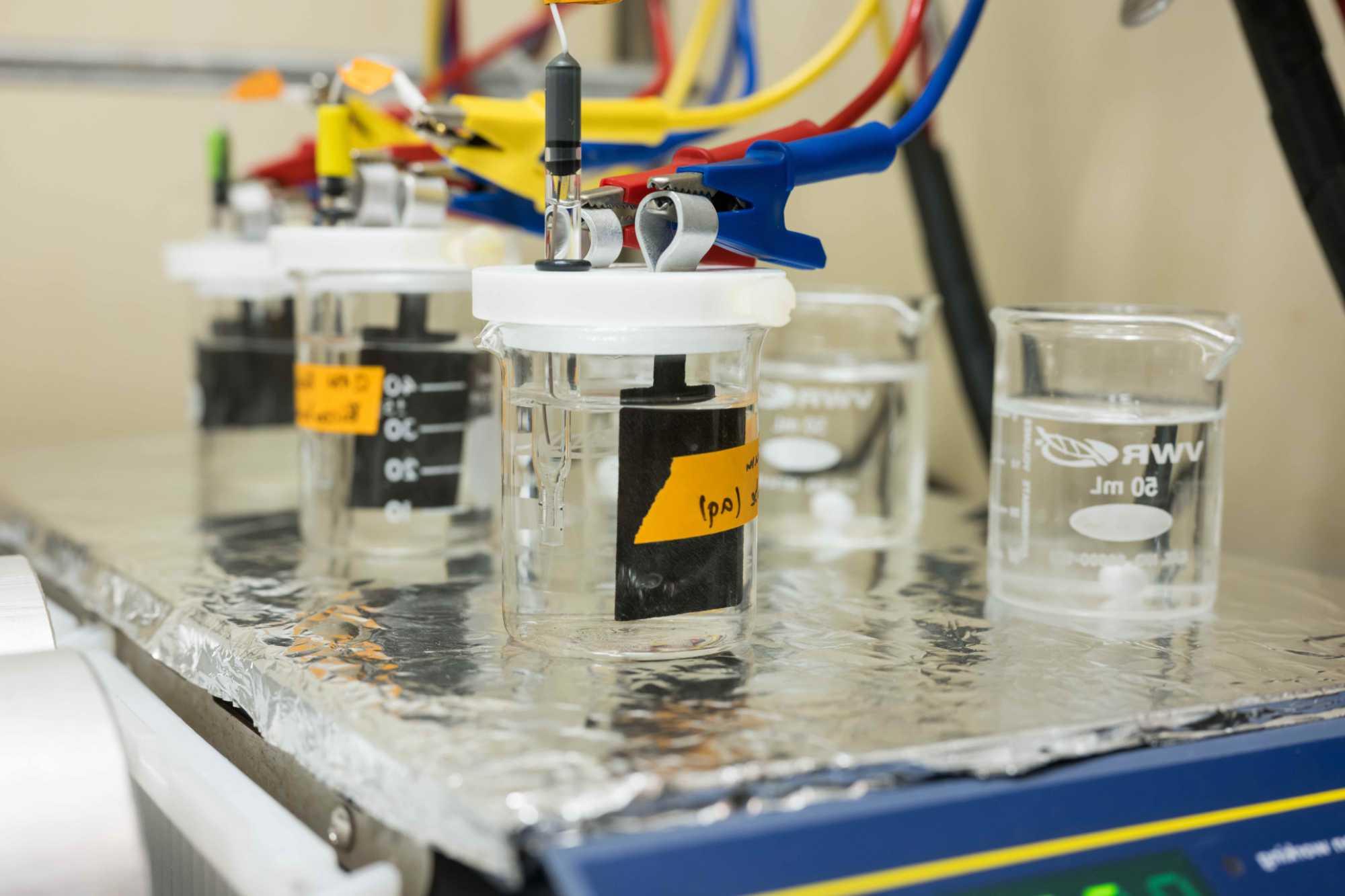 PFAS chemical remediation depicted with five beakers filled with water and hydrophilic carbon paper with blue, red, and yellow wires and jumper cable-style clamps affixed to them, all atop a scale covered with aluminum foil.